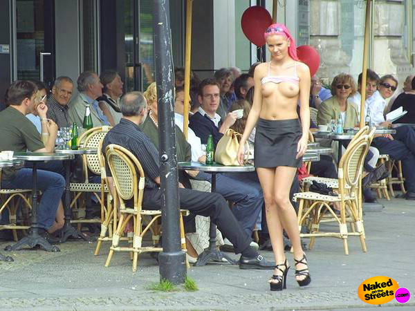Slender topless chick with pink hair walking by a crowded terrace